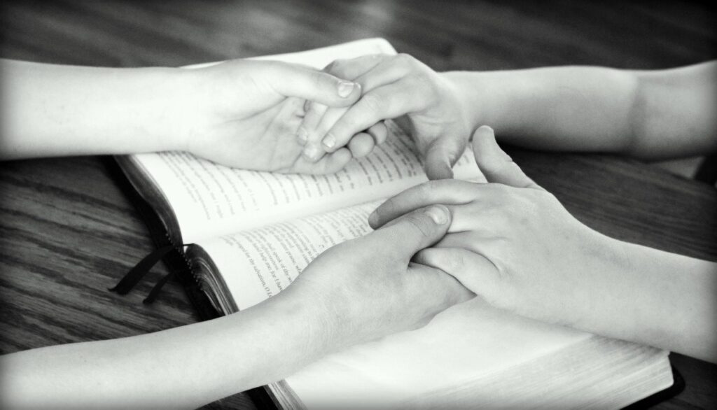 Black and white image of holding hands over the Bible praying. Being in the Word is pleasing to God.