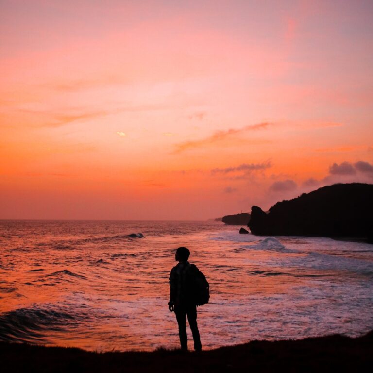 A person standing in front of a body of water with a pinkish orange sunset hue from Pexels.
